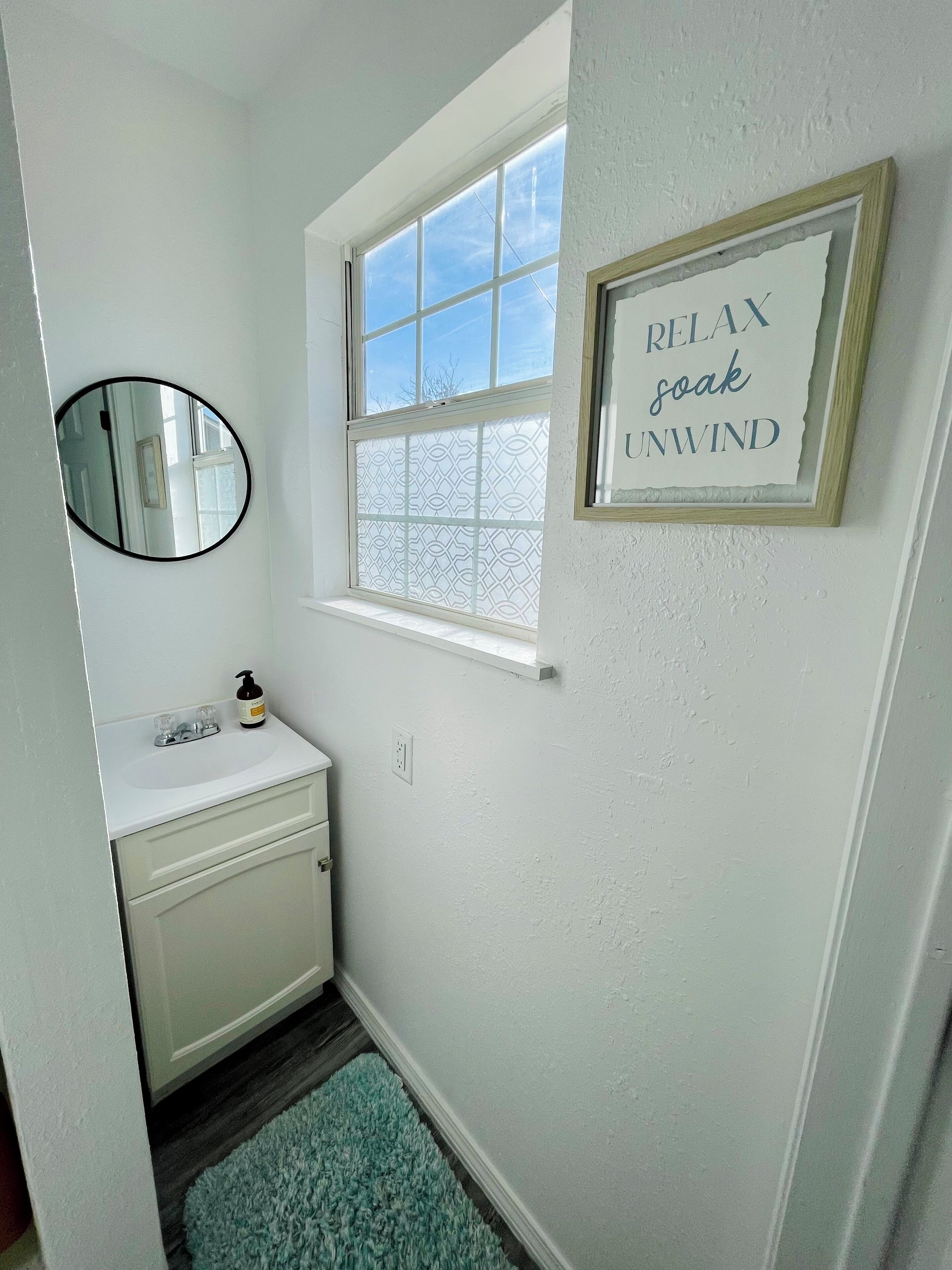 A bathroom with a white sink, a mirror, a window with a view, and a framed sign on the wall.