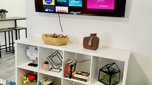 Una sala de estar con un televisor de pantalla plana montado en la pared que muestra varios servicios de transmisión. Debajo del televisor, hay un mueble blanco con estantes que contienen libros, objetos decorativos y un jarrón rojo.