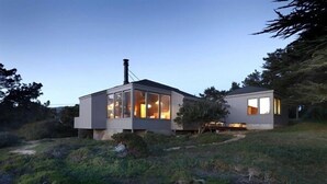 Et moderne hus med store vinduer og skorsten, omgivet af naturlig vegetation.