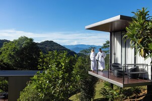 Un balcón moderno al aire libre con dos personas vestidas de blanco, con vistas a un paisaje exuberante y montañas a lo lejos.