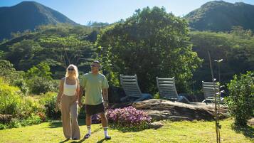Una pareja caminando en un jardín con montañas al fondo, sillones de descanso y una formación rocosa.