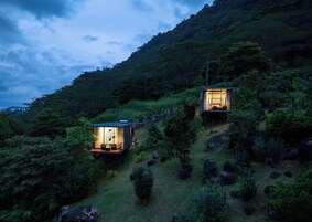 Dos cabañas modernas y elevadas ubicadas en un paisaje montañoso exuberante. Cada cabaña tiene un frente de vidrio, que permite vistas despejadas de la vegetación circundante. Las cabañas están conectadas por un sendero estrecho y sinuoso.