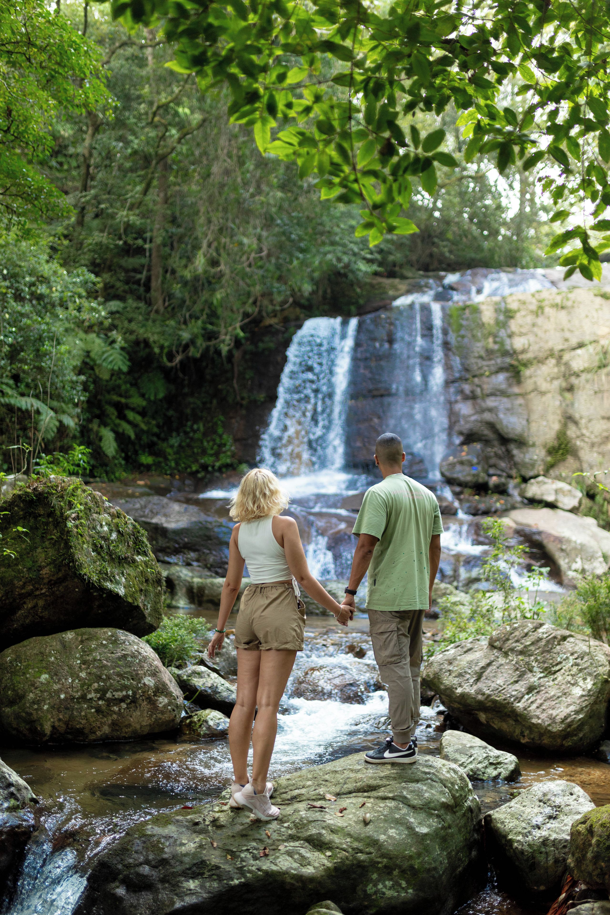 Two people standing on rocks by a waterfall in a forested area.