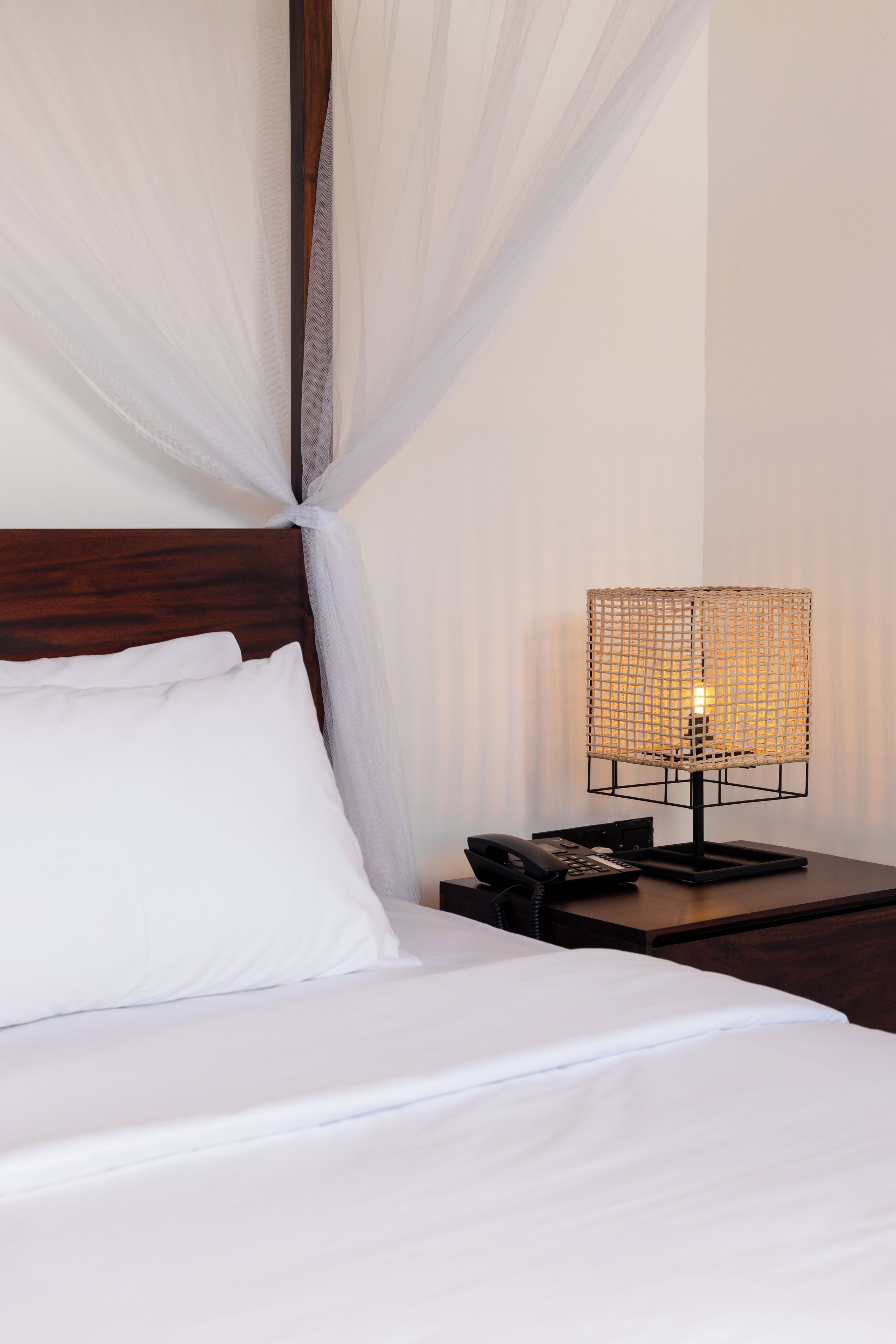 Un lit bien fait, avec du linge de lit blanc et une tête de lit en bois, une table de chevet avec une lampe et un téléphone.