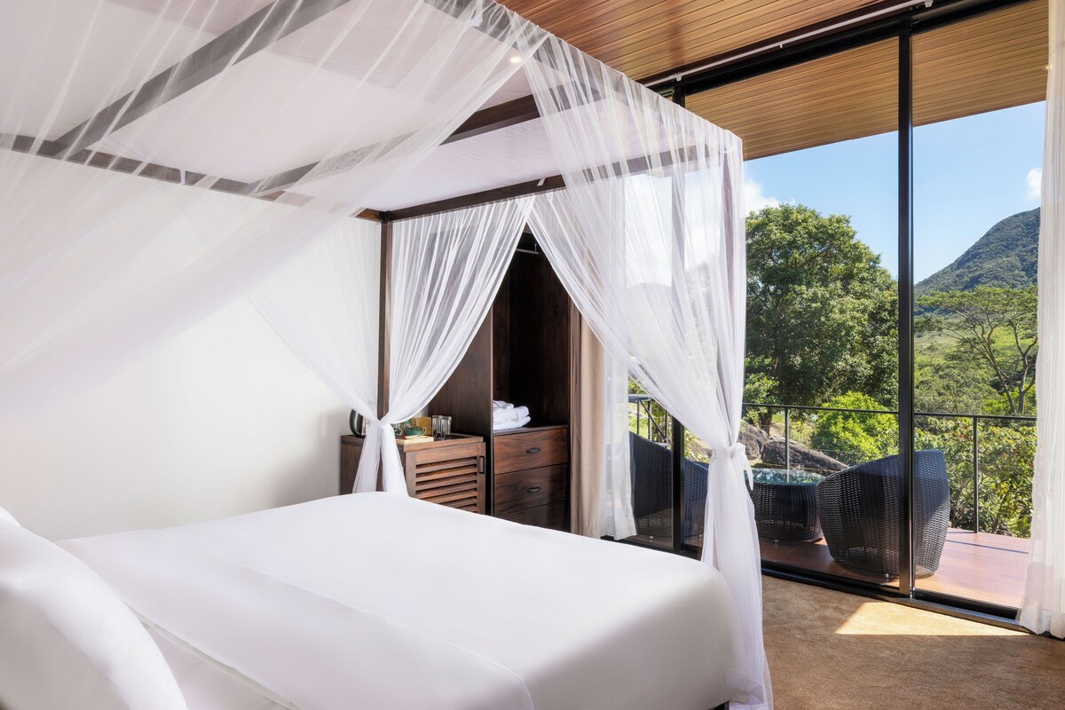 A bedroom with a canopy bed, a wooden dresser, and a view of a pool and mountains.