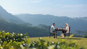 Un couple est assis à une table en plein air, profitant d’une vue panoramique sur des collines et des arbres en arrière-plan.