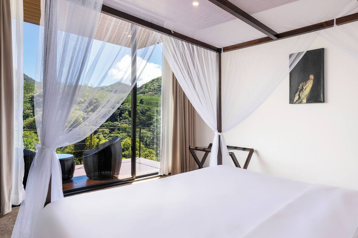 Un dormitorio con una cama grande, vistas a exuberante follaje y un balcón con mobiliario de mimbre.