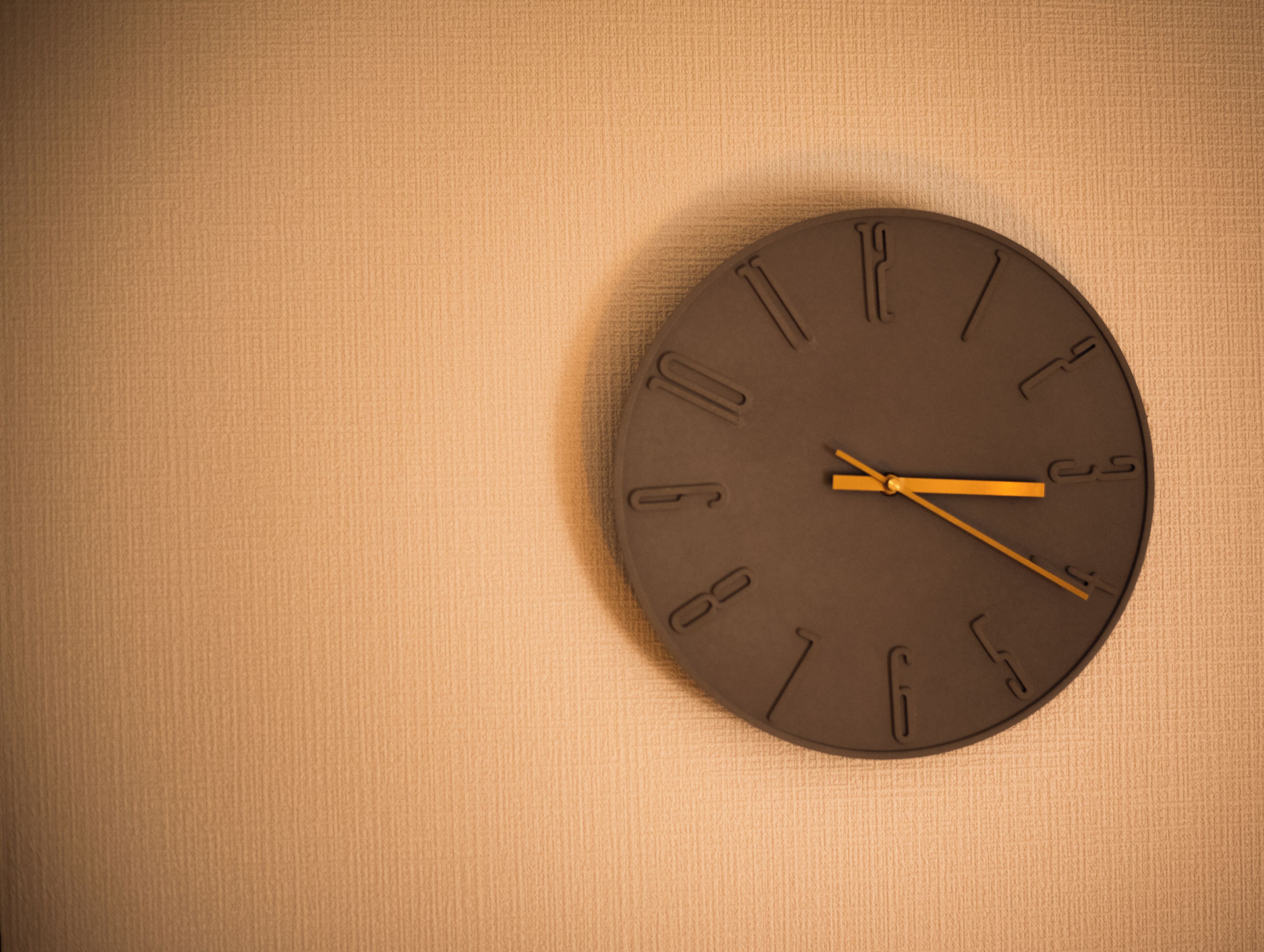A round wall clock with a brown face and yellow hands, mounted on a textured beige wall.