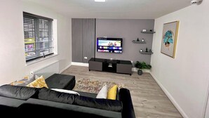 Un salon moderne doté d’un téléviseur à écran plat, d’un canapé noir, d’un tapis à motifs et d’une fenêtre avec des stores.