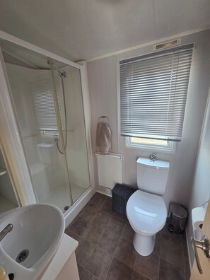 Un baño con ducha envuelta en vidrio, inodoro blanco, lavamanos y ventana con persianas.