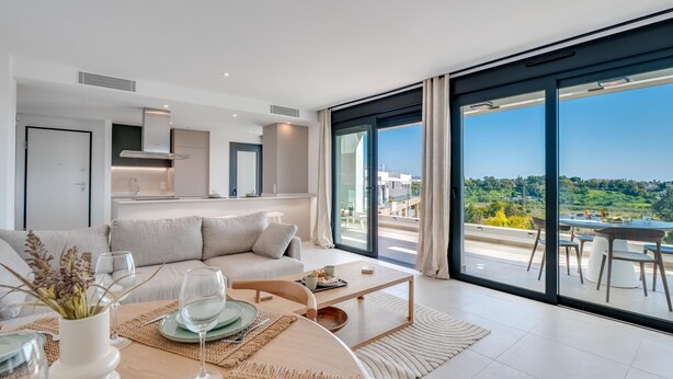 A modern living room with a dining area, large windows, and a view of the countryside. - Newly Built 2 BDRM Modern Oasis w Gym & Pool (Estepona)
