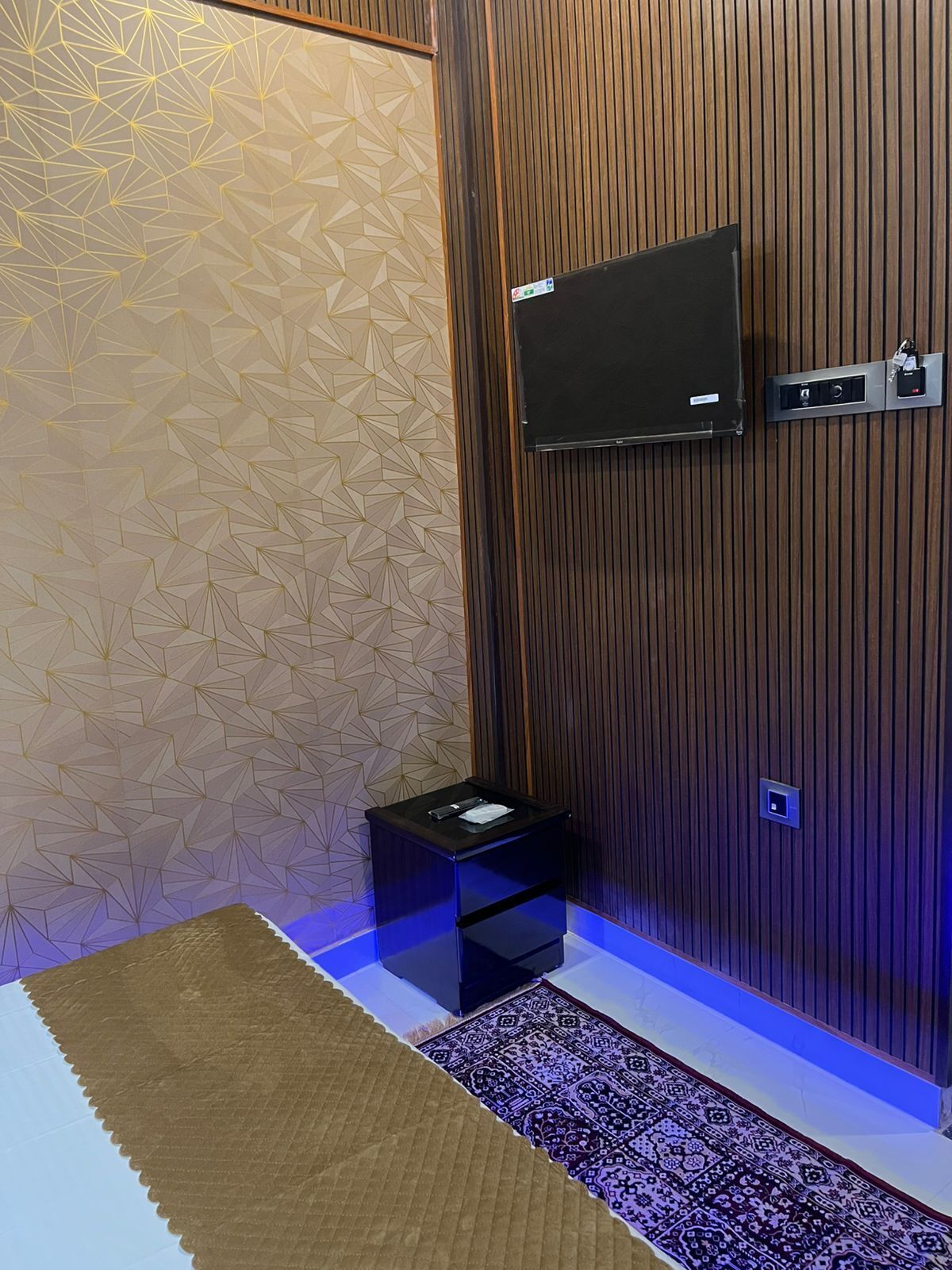 A modern bathroom with a geometric patterned wall, a mounted TV, a small black cabinet, and a patterned rug.