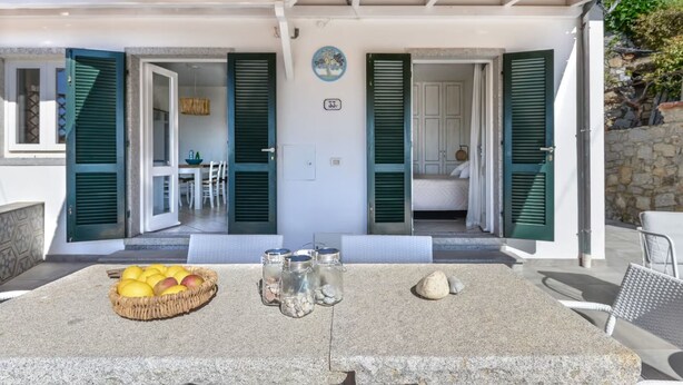 Un espace extérieur abrité avec une table en pierre, un panier de fruits, des salières et des poivrières, et une vue sur une pièce où l’on aperçoit un lit et une personne à travers une porte ouverte.