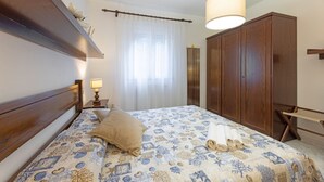 Un dormitorio con cama, armario, mesita de noche, lámpara y ventana con cortinas.