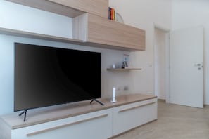 Ett modernt vardagsrum med en platt-TV på ett träställ, ett vitt skåp med lådor, en liten hylla med böcker och en klocka samt en vit dörr.