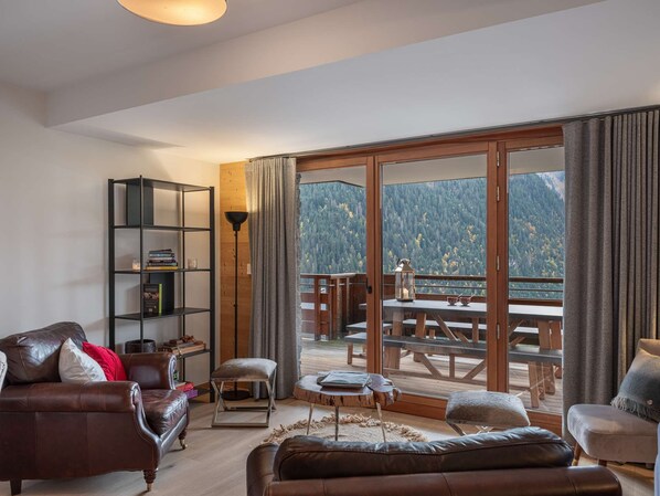 A modern living room with a leather sofa, a bookshelf, and a dining area with a view of a forested mountain. - Flambeaux B11 (CHATEL)