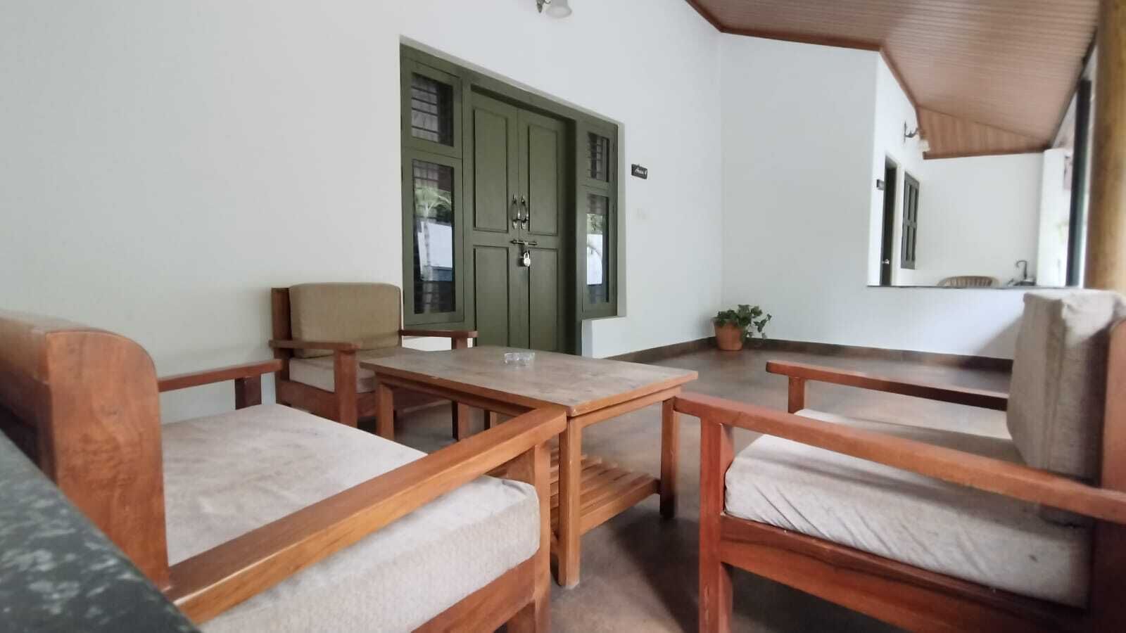 Een kamer met houten meubels, waaronder een bank, twee stoelen en een tafel. Er is een groene deur en een kleine potplant.