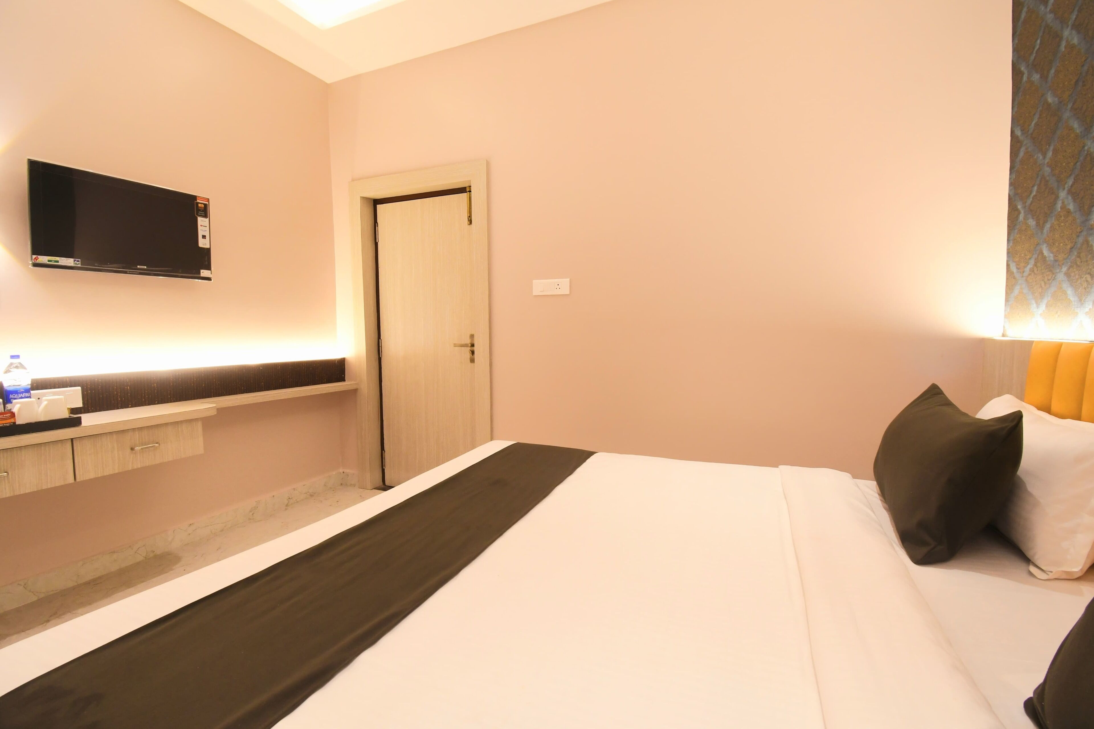 A hotel room with a bed, a TV mounted on the wall, a door, and a small counter with a bottle and a cup.