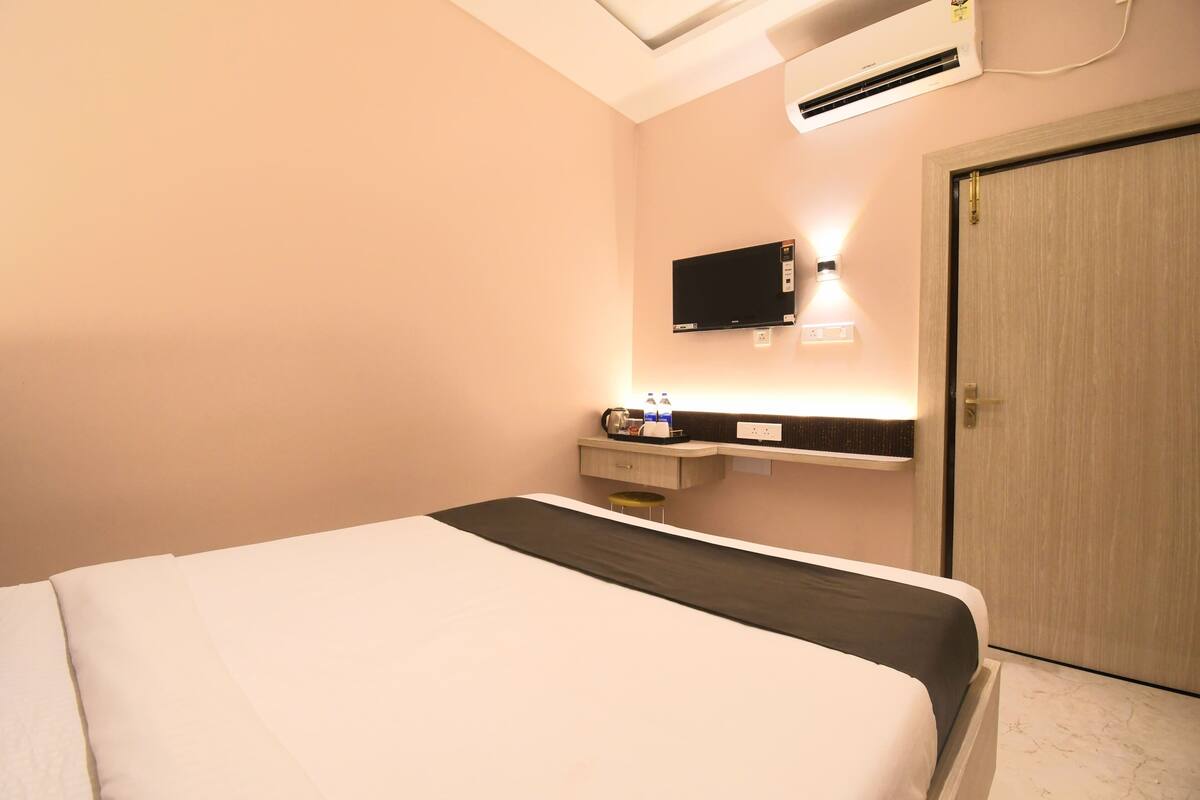 A hotel room with a bed, a TV mounted on the wall, a bedside table with a lamp, and a door.