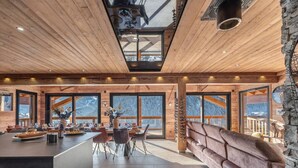 Une pièce spacieuse avec un plafond en bois, de grandes fenêtres offrant une vue sur les montagnes, un coin repas central avec une longue table et des chaises, et un mur d’accent en pierre.
