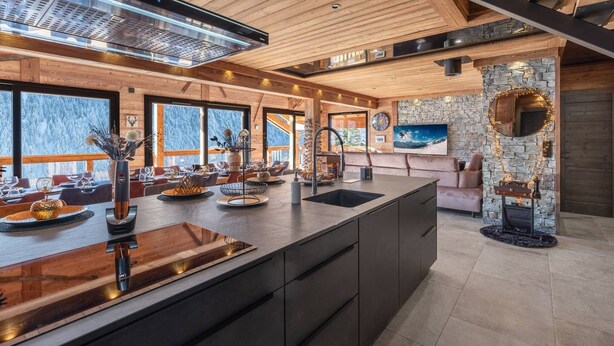 Une cuisine moderne dotée d’un grand îlot central, d’un plafond en bois, d’éléments décoratifs en pierre et de grandes fenêtres offrant une vue sur un paysage enneigé.