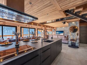 A modern kitchen with a large island, wooden ceiling, stone accents, and large windows offering views of a snowy landscape.