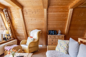 A cozy log cabin interior with a wicker sofa, a chair with a fur throw, a wooden dresser, and a radio. - Holiday Home 'Alwin' with Private Terrace and Wi-Fi (Goldlauter-Heidersbach)