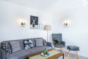 Uma sala de estar moderna com um sofá cinzento, almofadas estampadas, uma mesa de centro de madeira, um cacto como peça central, uma fotografia de um veado emoldurada e iluminação fixada na parede.