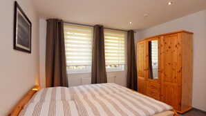 A bedroom with a bed, a wooden wardrobe, a window with blinds, and a framed picture on the wall.