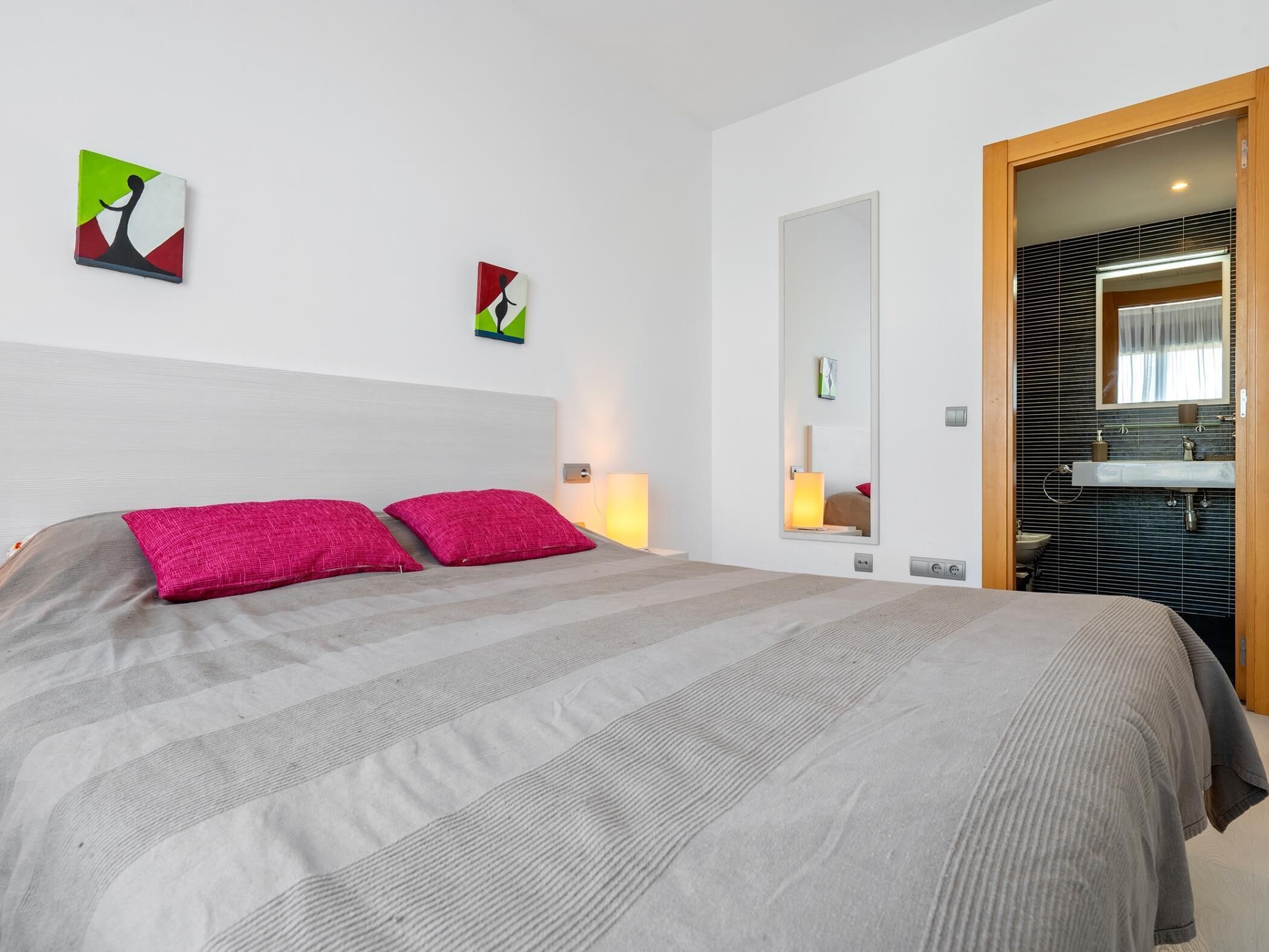 Une chambre d’hôtel avec un lit, une table de chevet avec une lampe, un miroir et une salle de bain visible à travers une porte ouverte.