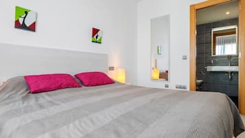 Une chambre d’hôtel avec un lit, une table de chevet avec une lampe, un miroir et une salle de bain visible à travers une porte ouverte.