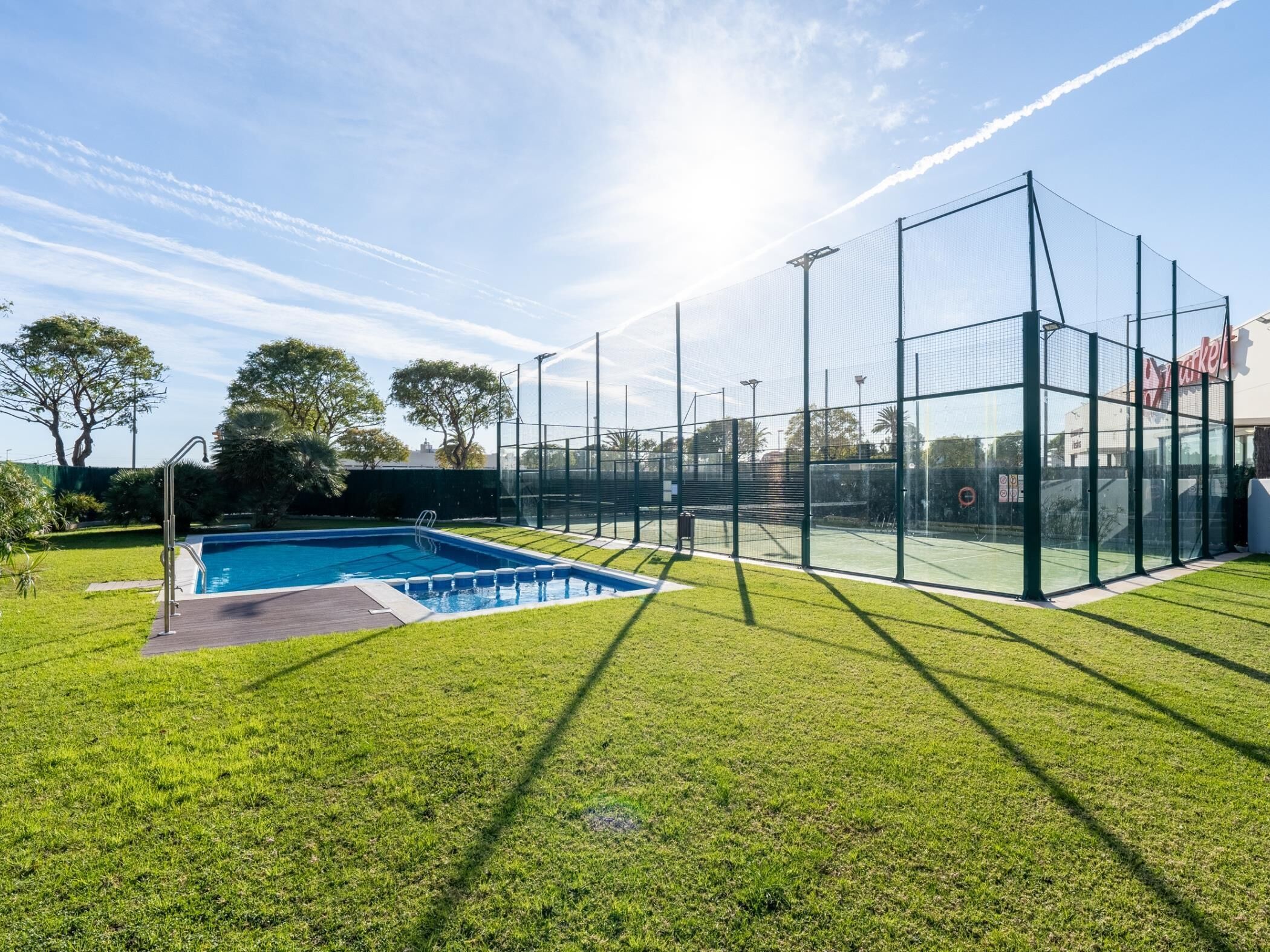 Una piscina con una valla alrededor, una cancha de tenis y un edificio con un letrero al fondo.