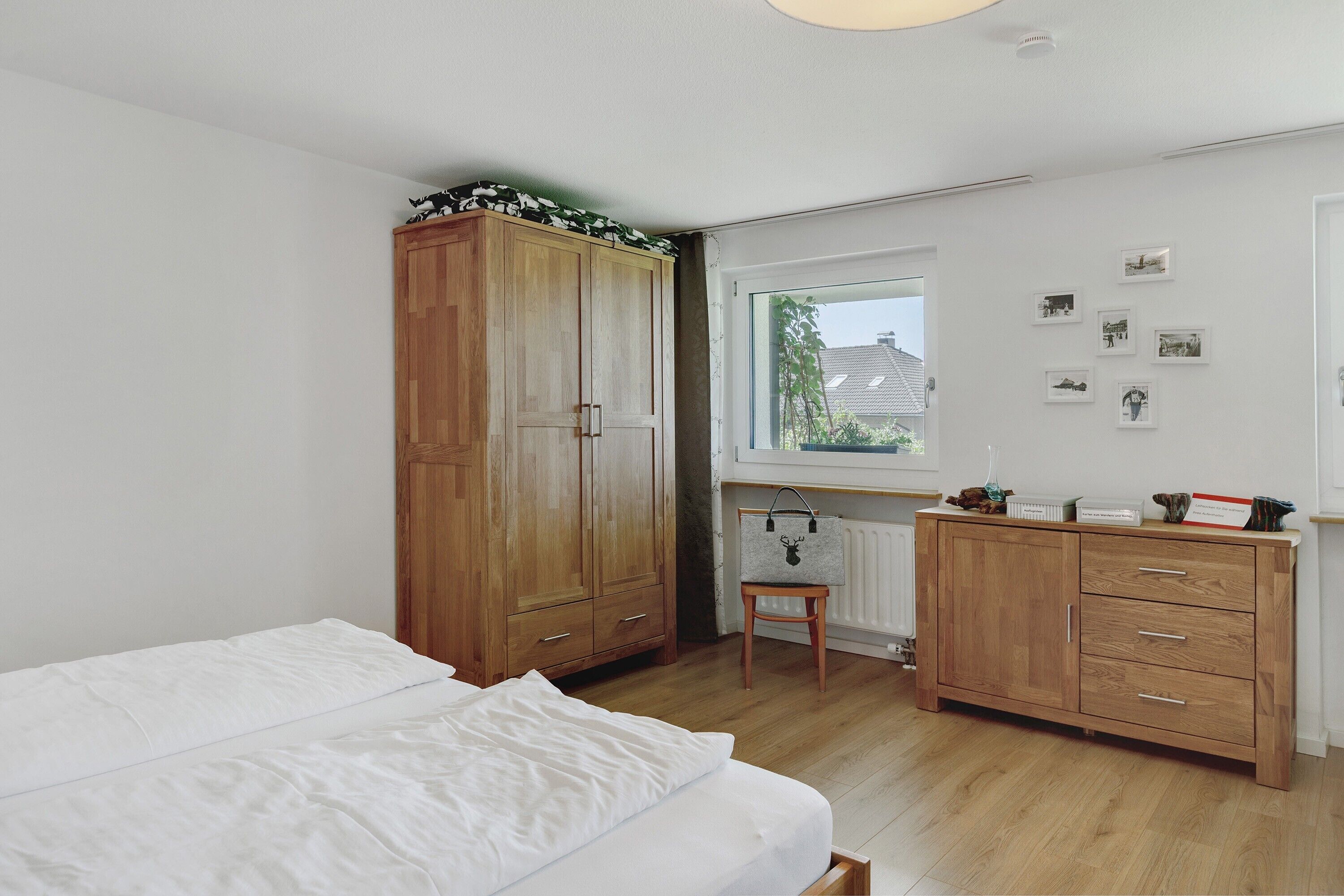 Ein Schlafzimmer mit Bett, Holzschrank, einer kleinen Bank, einem Heizkörper und einem Fenster mit Bildern an der Wand.