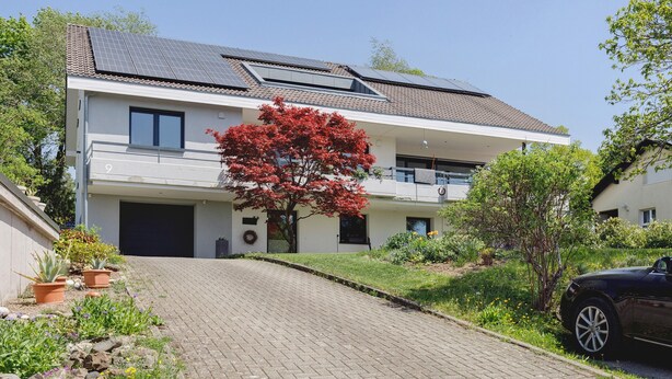 Et moderne hus med to etager, solpaneler, en asfalteret indkørsel og et rødt træ.