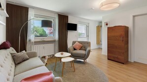 En moderne stue med sofa, lænestol, rundt bord og garderobeskab i træ.