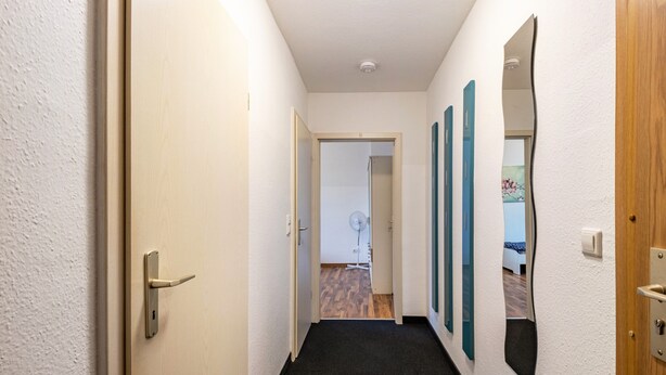 A narrow hallway with a carpeted floor, a wooden door on the left, and a mirror on the right. - Apartment 'Auf Zeit' with Wi-Fi (Leipzig)