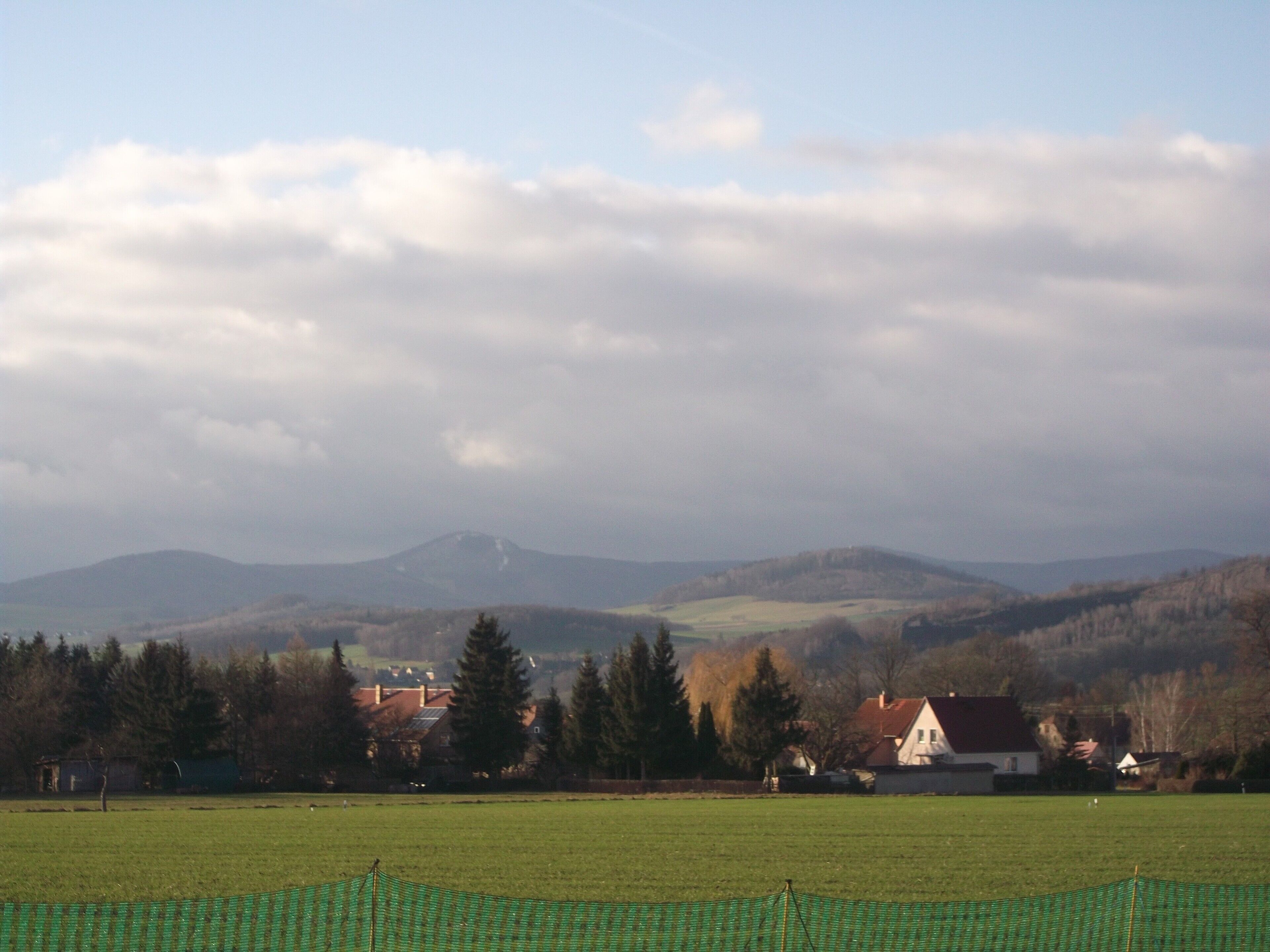 Un paysage rural avec une montagne à l’horizon, des maisons et un champ verdoyant.
