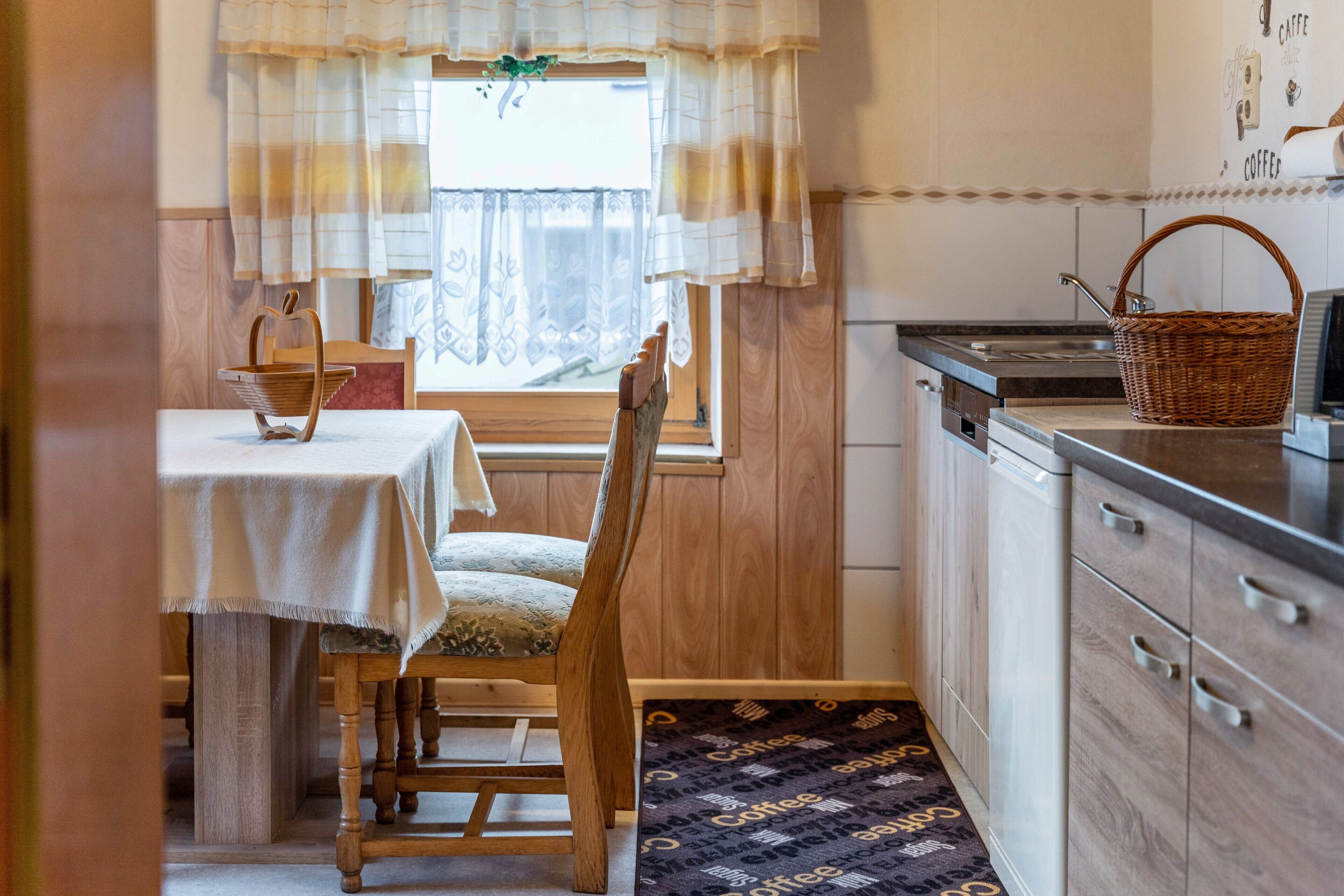 A kitchen with a dining table, wooden chairs, a wicker basket, and a window with curtains.