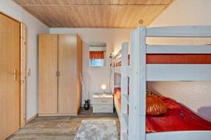 Ein Schlafzimmer mit Etagenbett, Holzschrank, Nachttisch mit Lampe und kleinem Fenster mit Vorhängen.