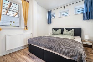 Une chambre à coucher comprenant un lit, une table de chevet, une fenêtre avec des rideaux, un radiateur et un plancher en bois.