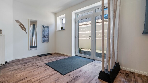 Ein Innenraum mit Holzboden, einer dunklen Matte im Eingangsbereich, einer Tür mit Glasfeldern und einer Wand mit Blattdekor.