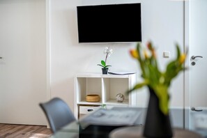 Een moderne kamer met een flatscreen televisie aan de muur, een klein wit schapje met een bloempot, en een zithoek met een glazen tafel en stoelen.