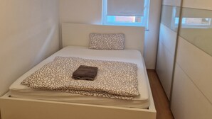 A small, neatly made bed with a patterned bedspread and a folded towel, a window with blinds, and a wooden floor. - Fantastic Apartment in the Heart of Wedel (Wedel)