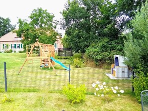 Un espace de jeux extérieur clôturé, comprenant une structure d’escalade en bois, un toboggan et un trampoline.