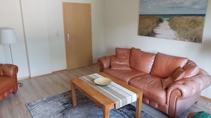A living room with a brown leather sofa, a wooden coffee table with a bowl, a lamp, and a painting of a beach.