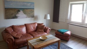 A living room with a brown leather sofa, a wooden coffee table, a small ottoman, a floor lamp, and a painting of a beach scene.