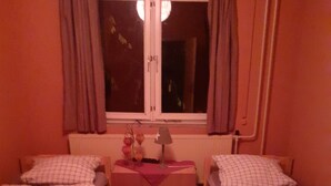 A small, dimly lit room with two single beds, a small table with a lamp, and a window with curtains.