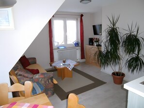 Un salon comprenant un canapé, une table basse, une table à manger, une plante en pot et un coin cuisine avec un téléviseur et des placards.