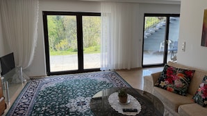 Un salon agrémenté d’un grand tapis à motifs, d’un canapé avec des coussins à fleurs, d’une table basse avec une télécommande, et offrant une vue sur un jardin grâce à de grandes portes coulissantes.