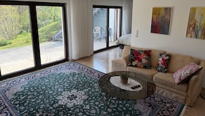 Un salon comprenant un canapé, une table basse en verre, un tapis à motifs et une vue sur un espace extérieur, accessible par de grandes portes vitrées.